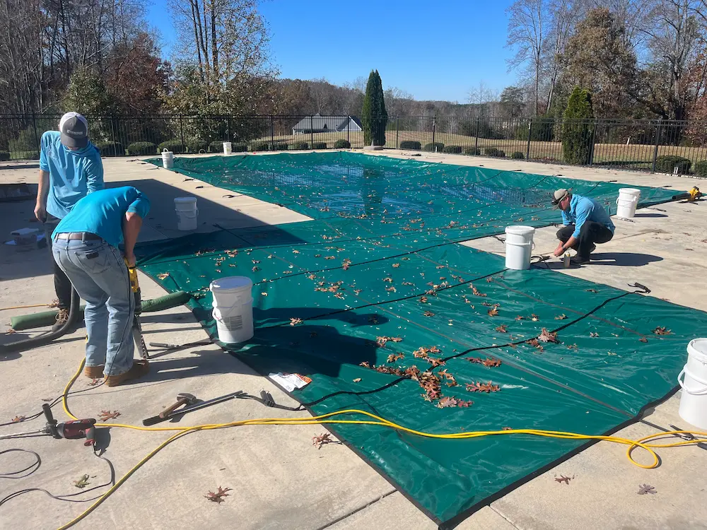 Technicians fitting a green mesh safety cover on an in-ground pool